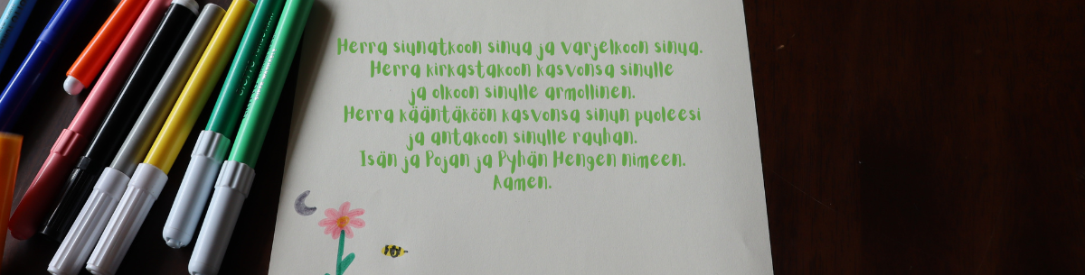 Herran siunaus kirjoitettuna paperille, jossa on piirretty kukka ja mehiläinen, sekä pöydällä tusseja.